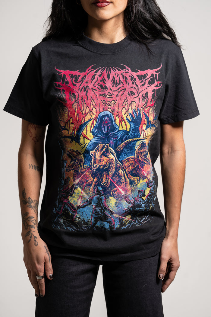 A person with long dark hair and tattooed arms wears a Galactic Empire black T-shirt with colorful artwork of a hooded figure and three fierce dinosaurs in a dynamic, fiery scene. The shirt displays a stylized band logo.