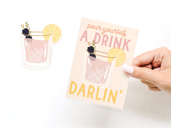 A hand holds Onderkast Studio’s Pour Yourself a Drink Darlin Cocktail Sticker Greeting Card, printed on white card stock with a cocktail design. A matching vinyl sticker of the drink is placed next to the card.