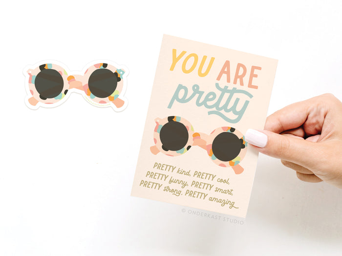 A hand holds the Onderkast Studio You Are Pretty Sunglasses Sticker Greeting Card, made of thick white stock with colorful sunglasses and bold text. A matching vinyl sticker with the same design is beside the card.